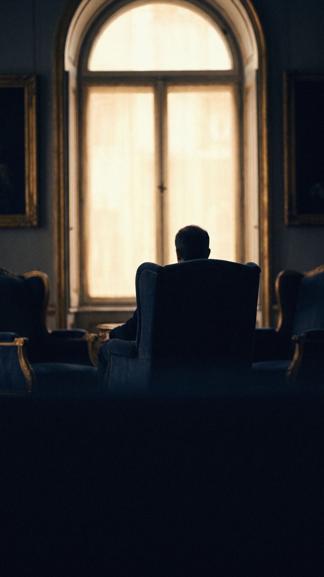 Un homme assis dans un fauteuil dans une pièce vaste et sombre.