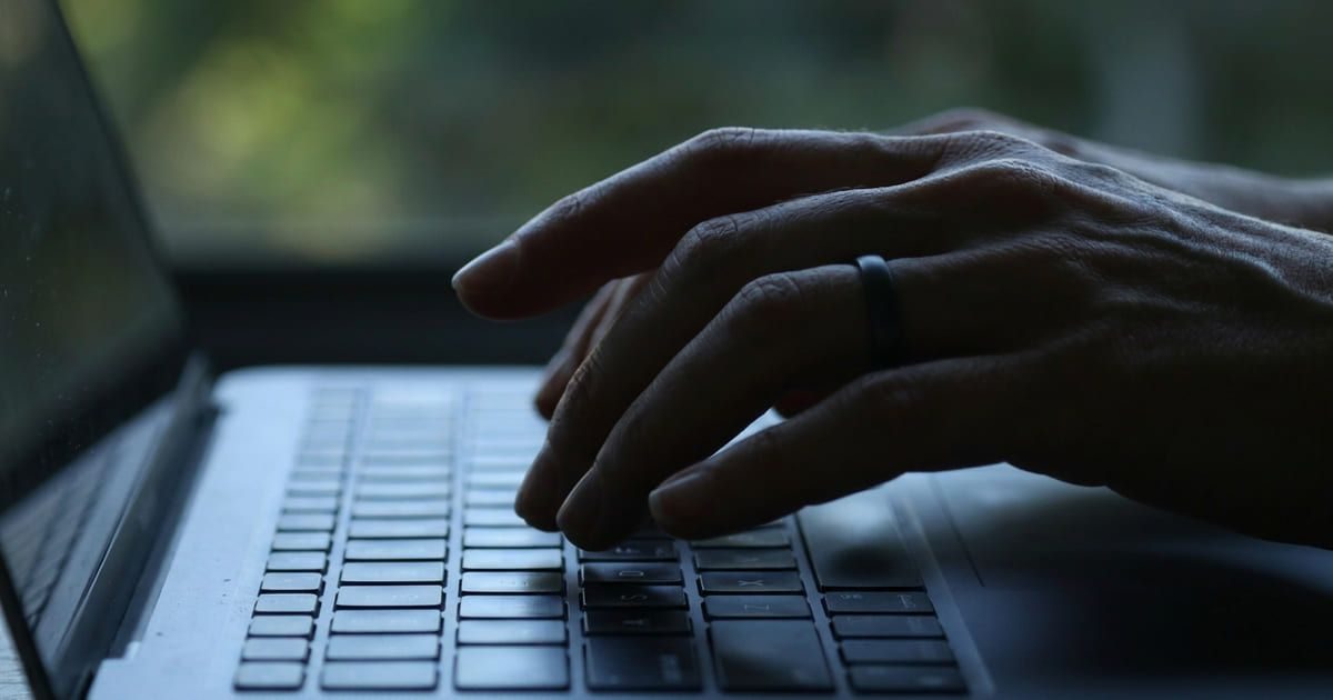 Mão a escrever no teclado de um portátil, em contraluz, junto a uma janela